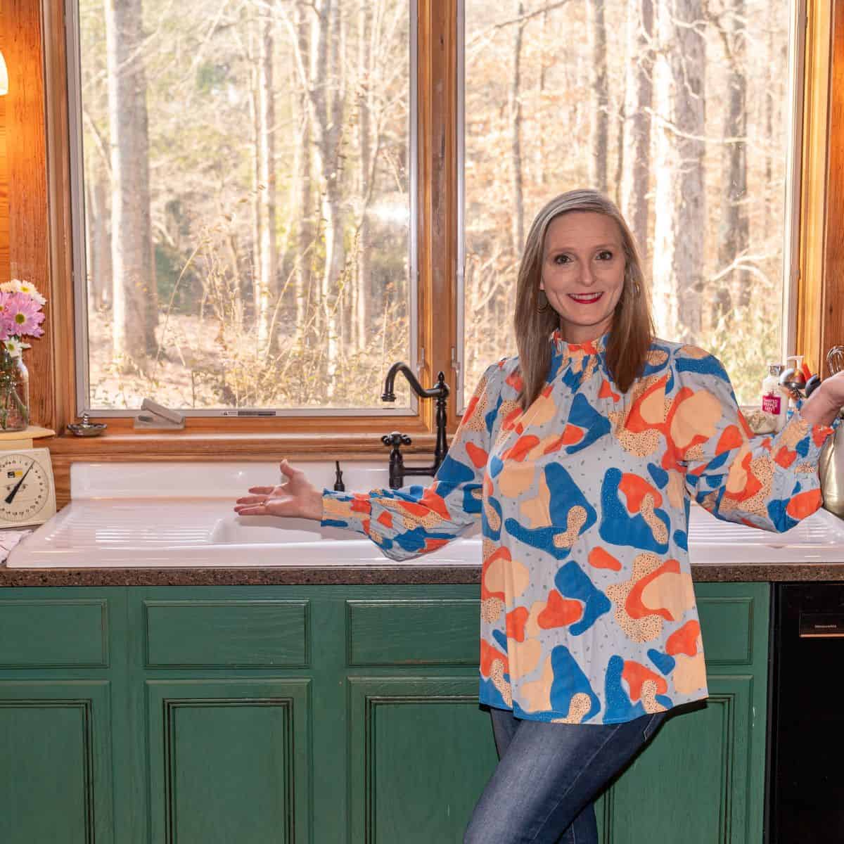A woman standing in front of her kitchen window by her sink.