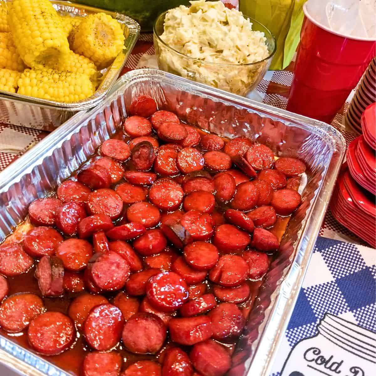 BBQ Sausage Bites in an aluminum pan. Corn on the cob, potato salad a red solo cup and blue and white table cloth on the table.