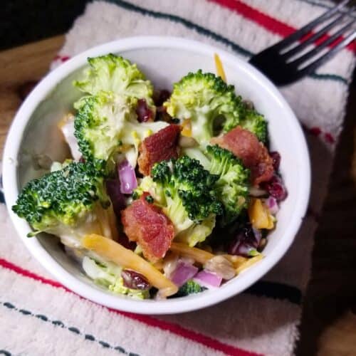 Bacon broccoli salad in a white bowl with a fork and a stripped dish towel.