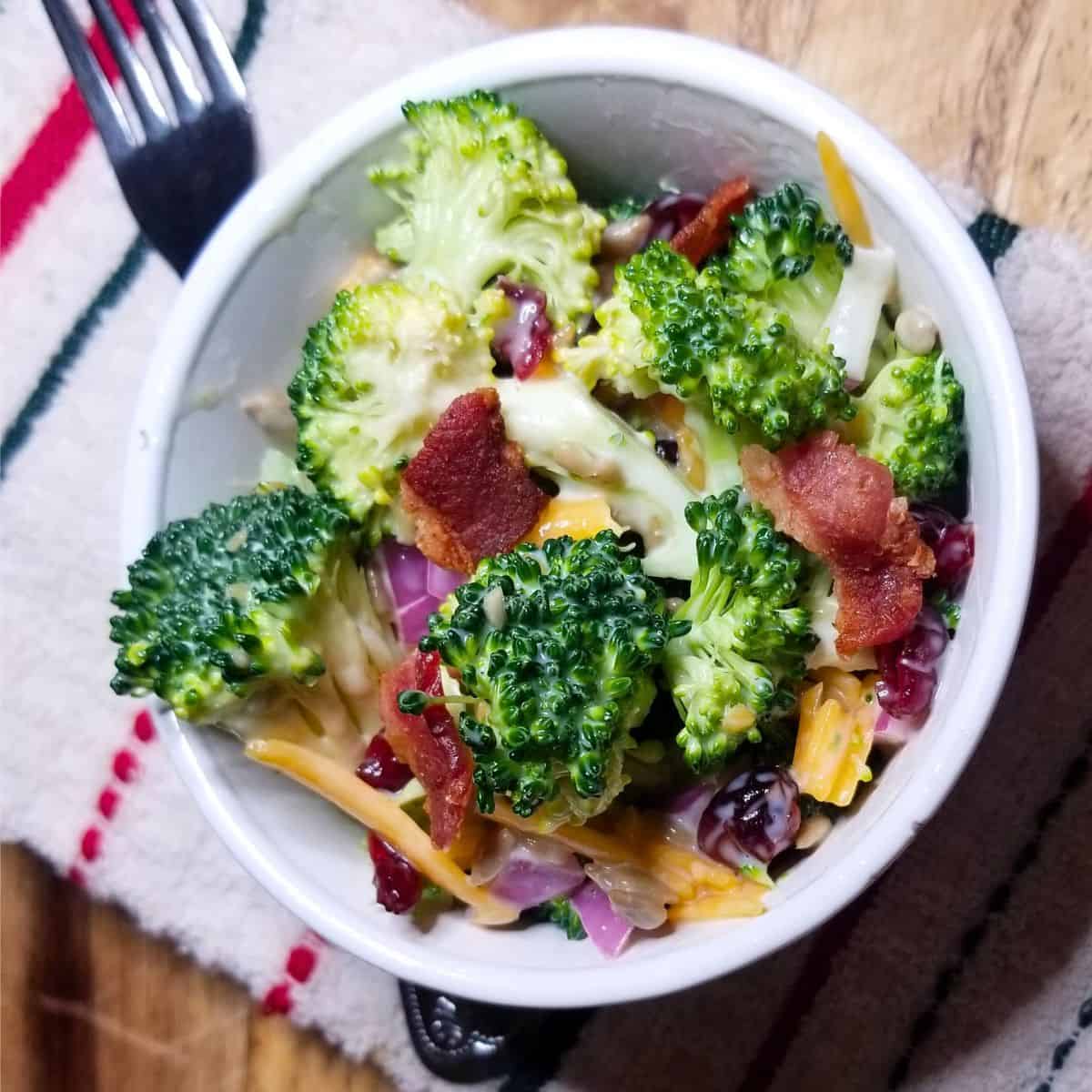 Bacon Broccoli salad in a white bowl with a fork and stripped dish towel.
