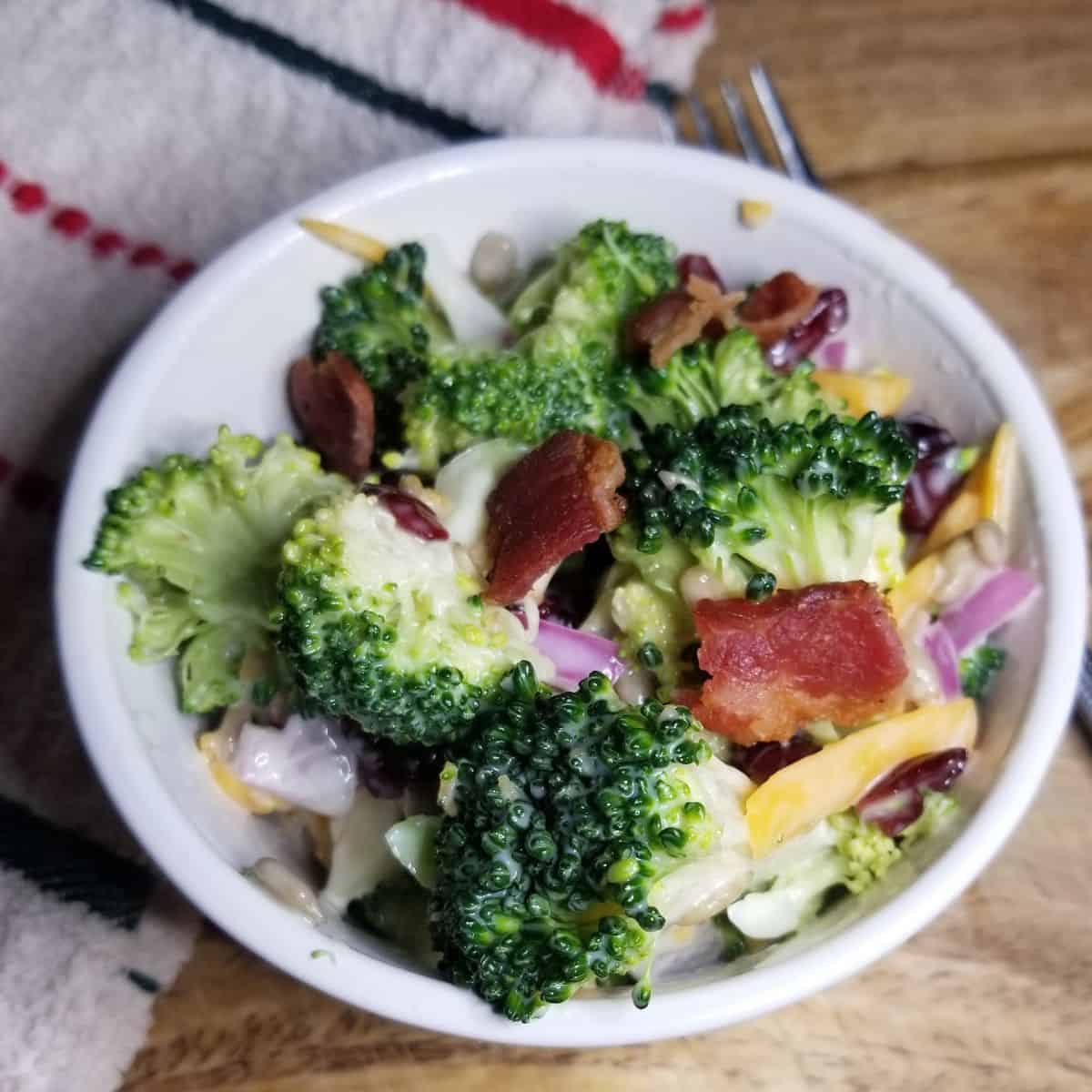 Broccoli salad in a white bowl with a fork a stripped towel next to the bowl.