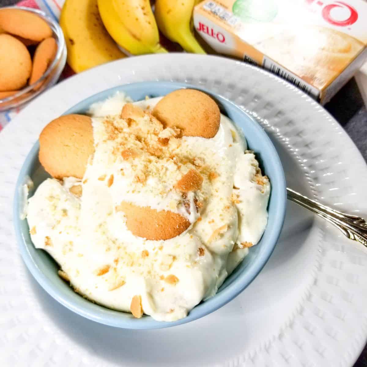 Banana pudding in a blue bowl sitting on a white plate with vanilla wafers, bananas, and instant pudding in the background.