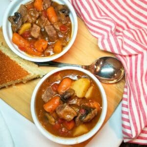 Beef Stew in two white bowl with a spoon and cornbread. A red and white dish towel next to the bowls.