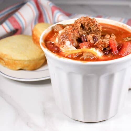 Chili in a white bowl with cornbread on a plate. Dish towel in the background.