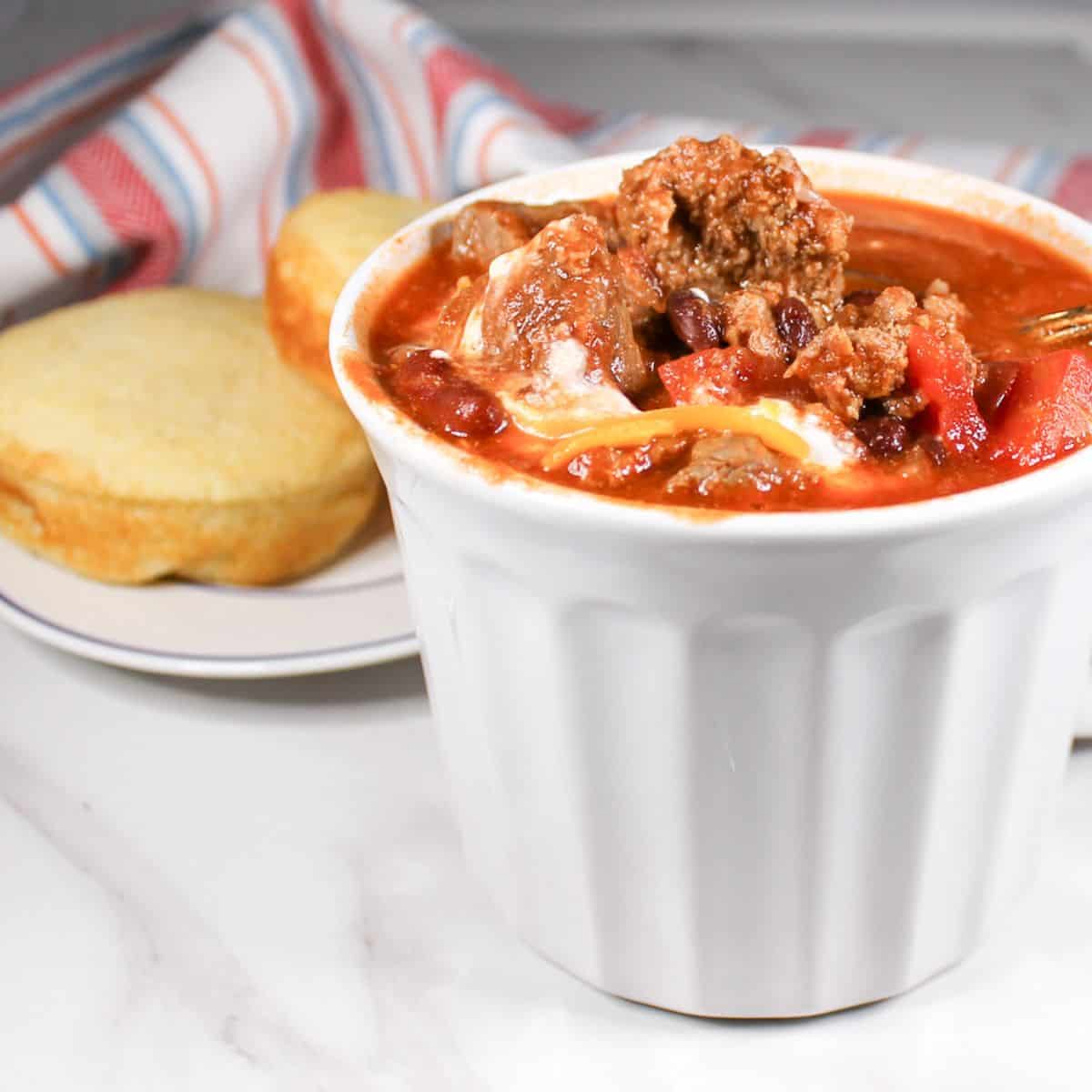 Chili in a white bowl with cornbread on a plate. Dish towel in the background.