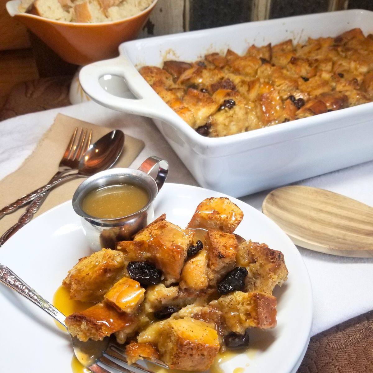 Freshly baked bread pudding with golden brown top in white baking dish and serving plate. Utensils on the side. Overhead view of bread pudding dessert on rustic table setting.