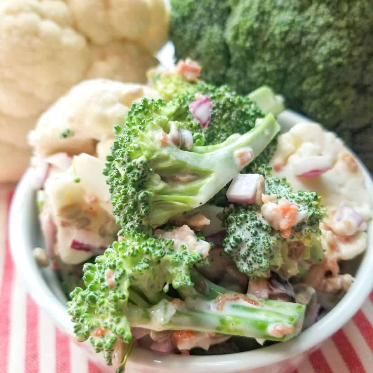Broccoli salad in a white bowl with vegetables in the background.