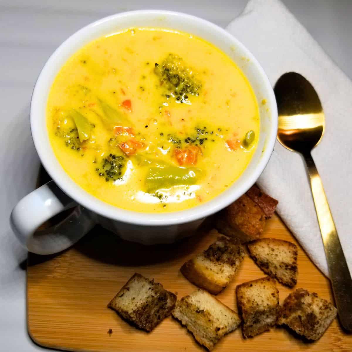 Broccoli Cheese Soup in a white bowl. A wooden cutting board under the bowl with a gold spoon, white napkin and croutons.