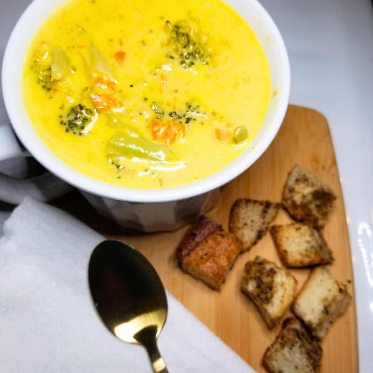 Broccoli Cheddar Soup in a white bowl with a gold spoon. Croutons and white napkin on a wooden board.