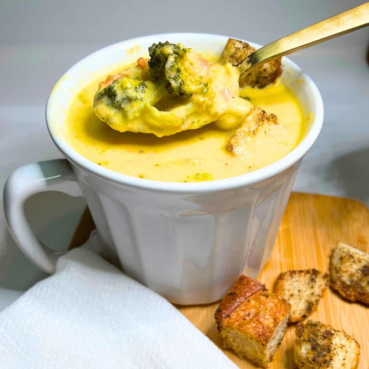 Broccoli cheese soup in a white bowl with a gold spoon. Croutons and a white napkin next to the bowl.