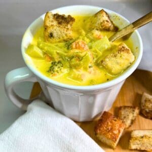 Broccoli cheddar soup in a white bowl with croutons in the soup and on a wooden tray. White napkin on the tray. Gold spoon in the bowl.