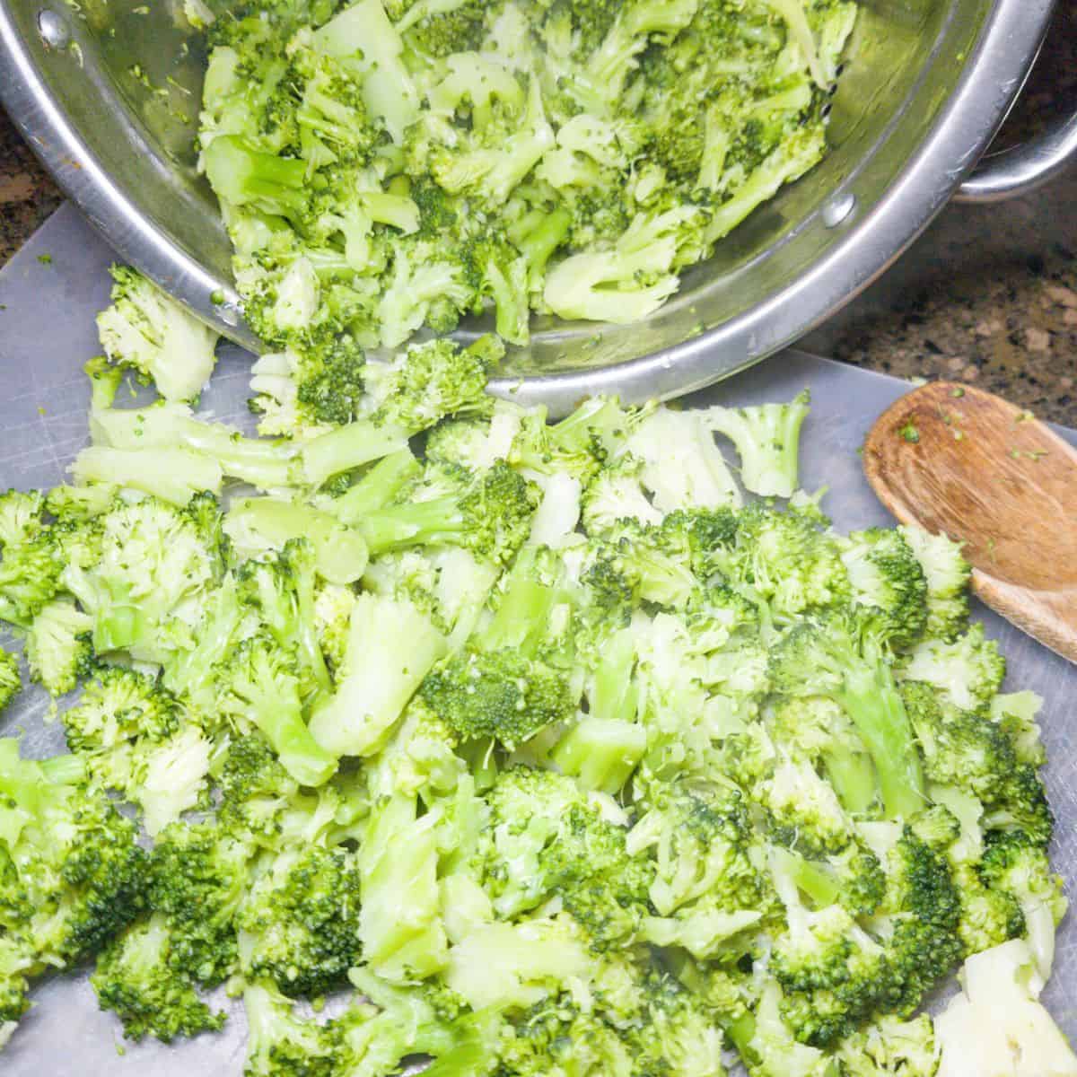 Cooked broccoli on a cutting board with more in a colander. A wooden spoon to the side.