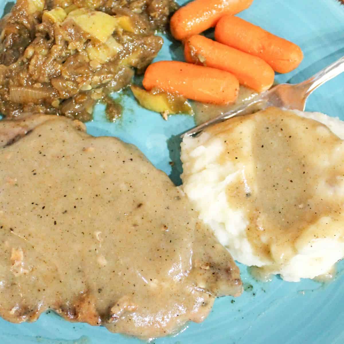 Brown gravy over a pork chop and mashed potatoes with other vegetables on a blue plate.