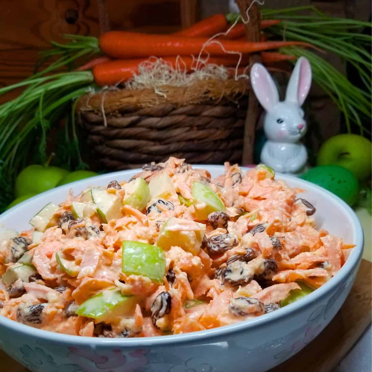 Carrot Apple Salad in a bowl with carrots sitting in a basket in the background. A glass rabbit and apples on the table.