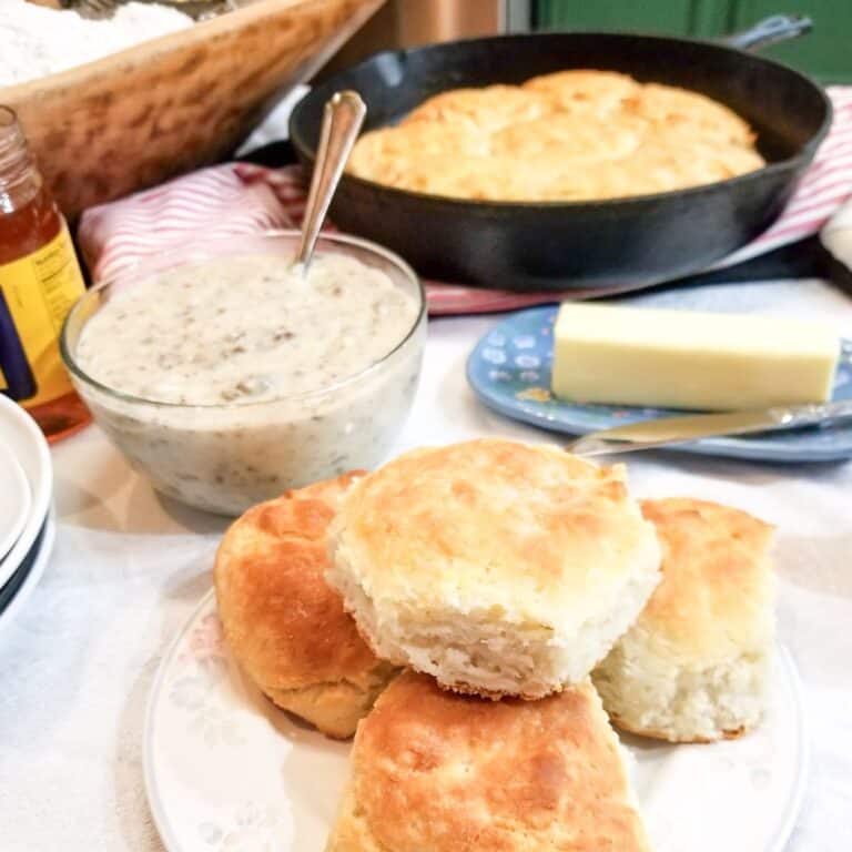 Cathead biscuits on a white plate with a bowl of gravy, butter, and a cast iron skillet of homemade cathead biscuits. A wooden bowl of flour and a jar of syrup in the background.