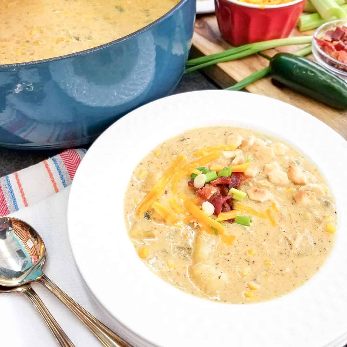 Chicken corn chowder served in a white bowl. A pot of chowder and ingredients in the background.