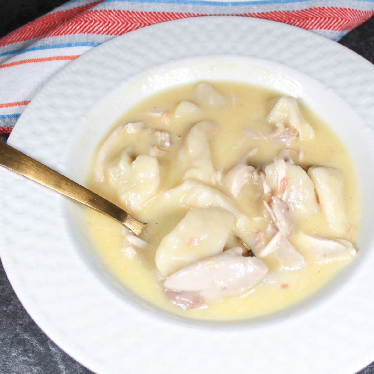 Chicken and Dumplings in a white bowl with a gold spoon. A stripped dish towel next to the bowl.