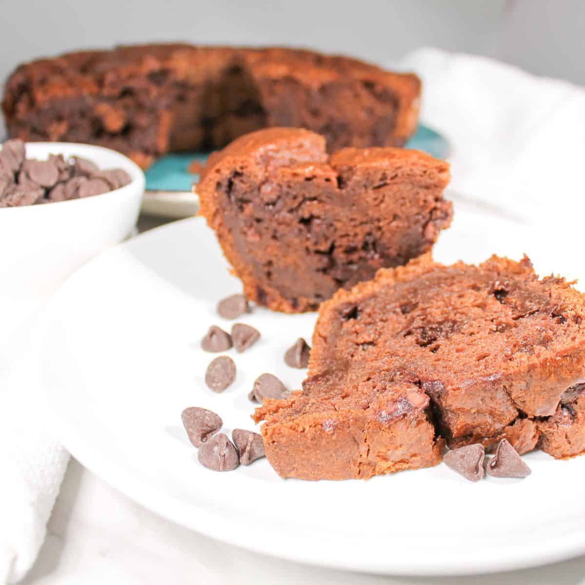 Sliced chocolate cake on a white plate with more cake in the background on a blue plate. Chocolate chips added to the sliced cake.