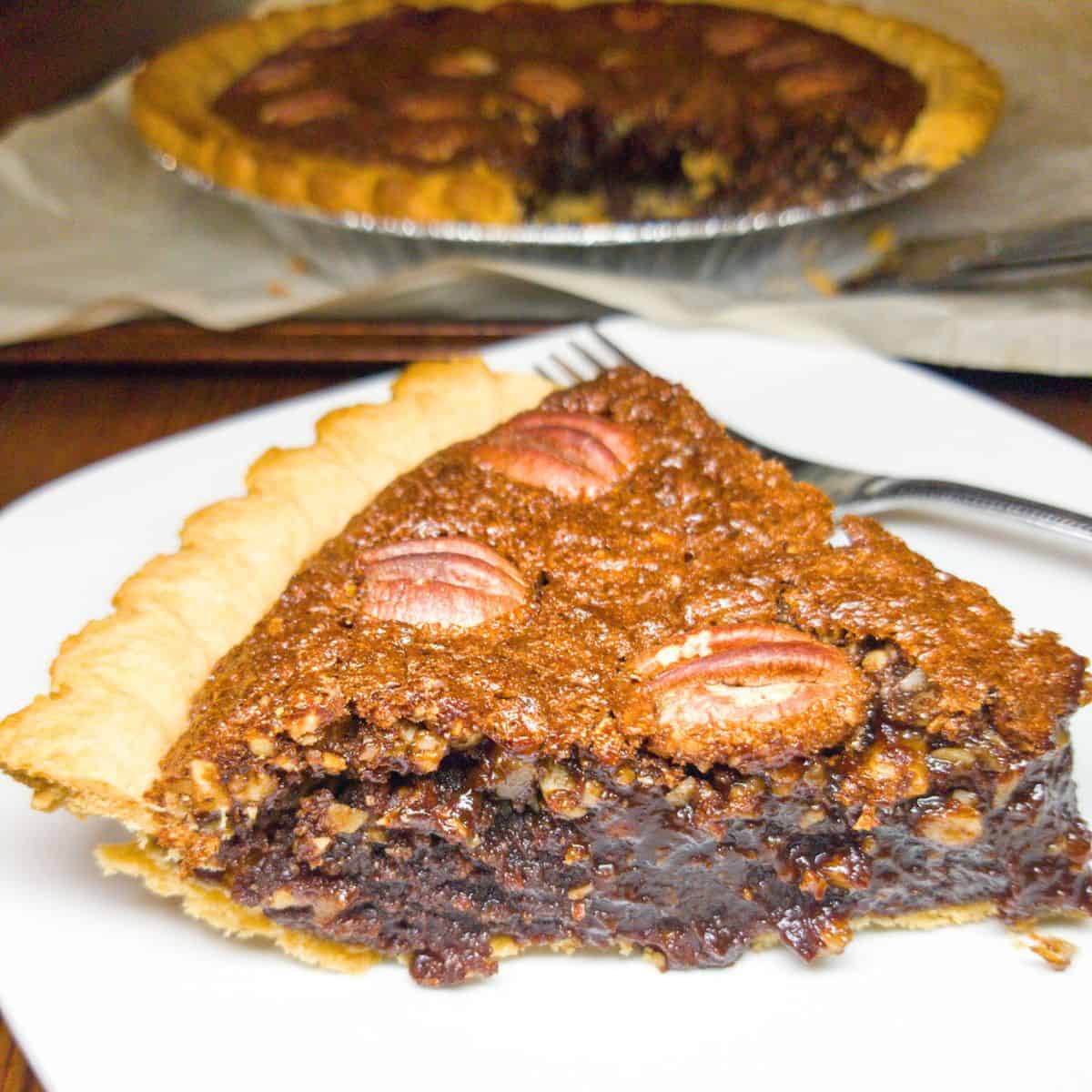 Chocolate pecan pie on a white plate with a fork and a whole pie in the background.