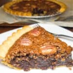 Chocolate pecan pie on a white plate with a fork and a pie in the background.