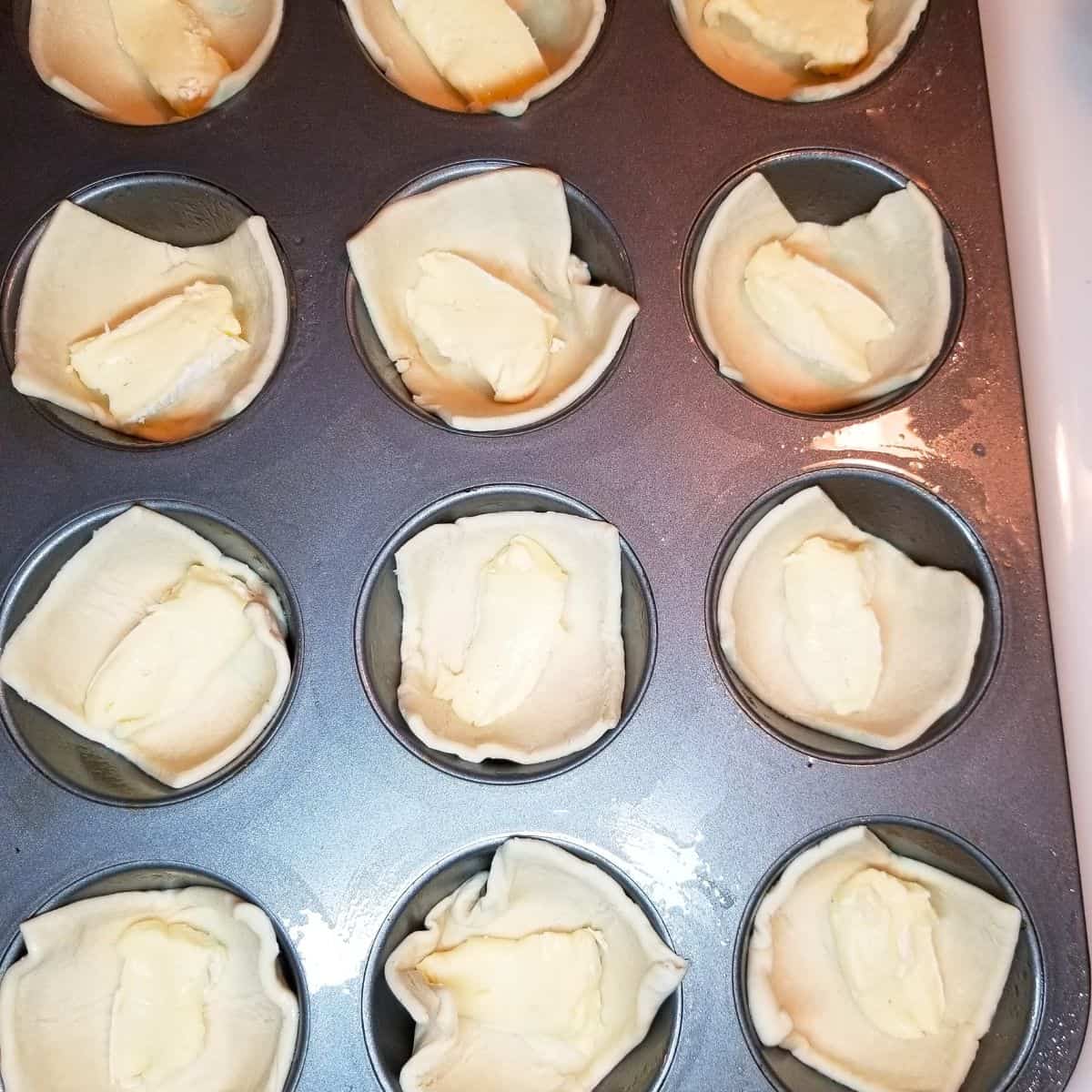 A muffin tin with puff pastry and brie cheese inside each well.