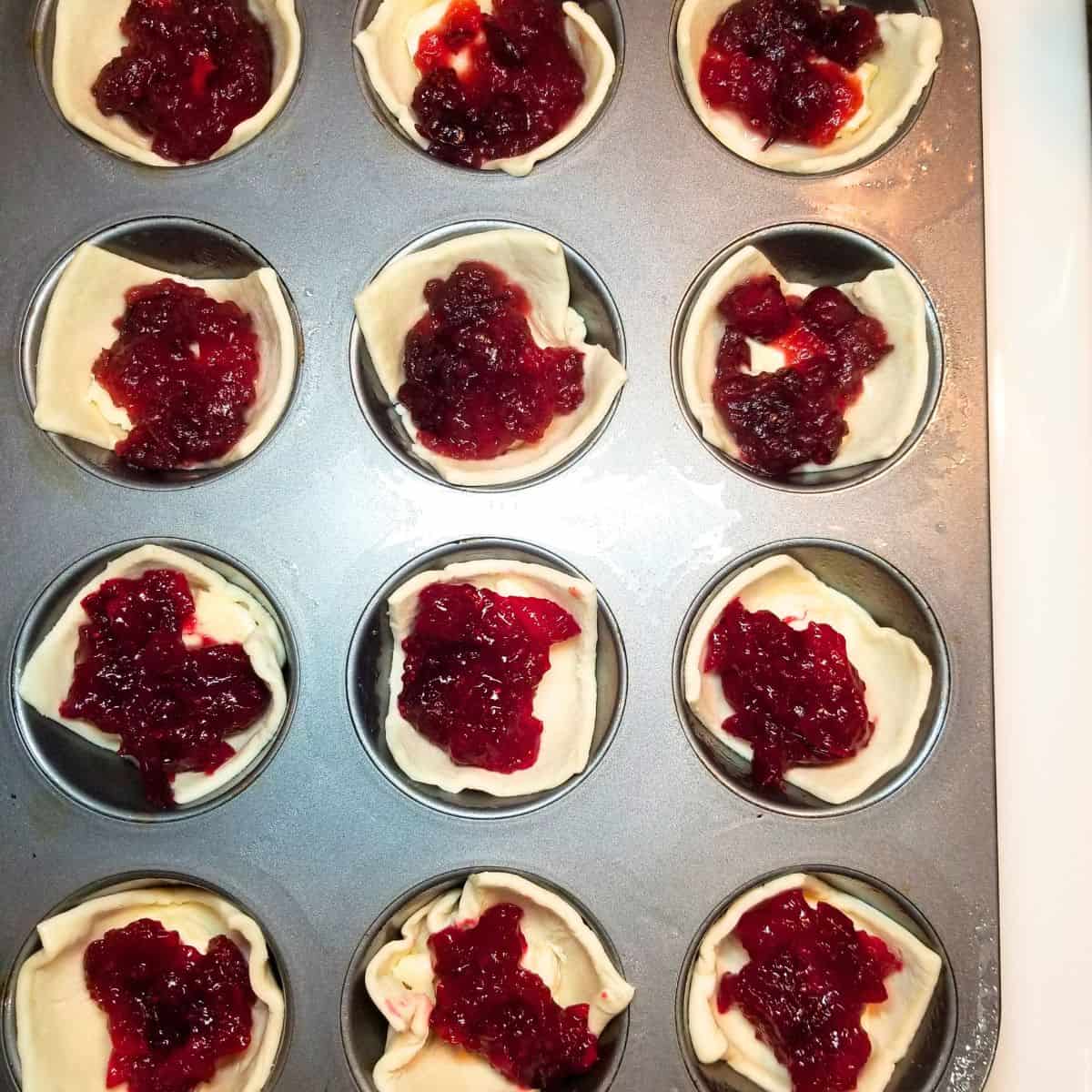 A muffin tin with puff pastry, cheese, and cranberry sauce inside each well.