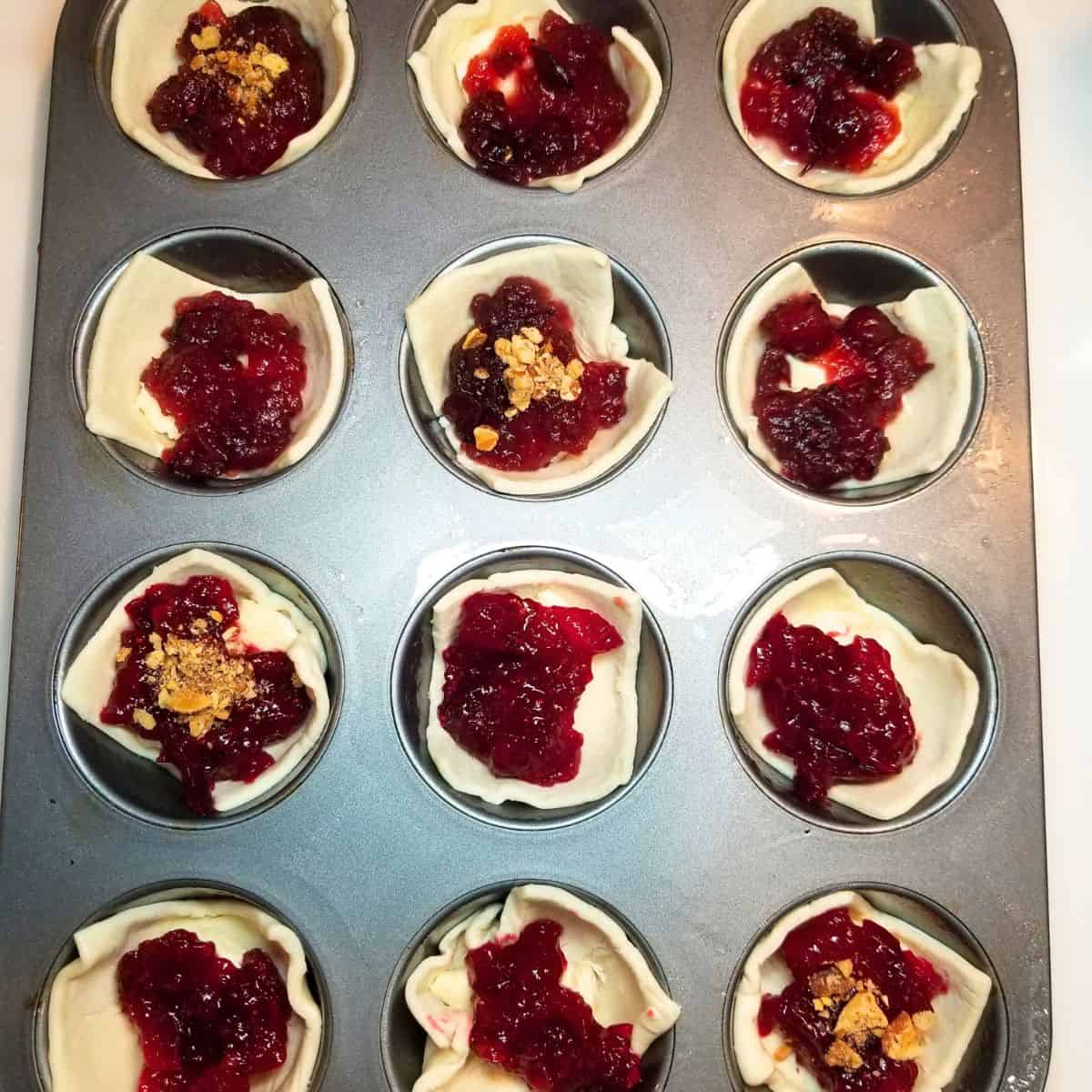 A muffin tin with puff pastry, cheese, and cranberry sauce inside each well. A few have chopped nuts.