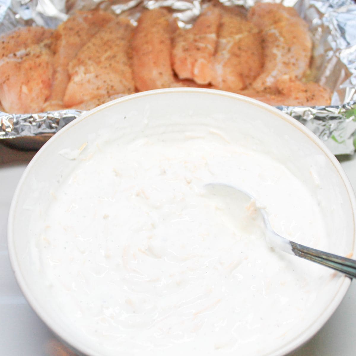 A white bowl with French onion dip mix and uncooked chicken tenders in the background.