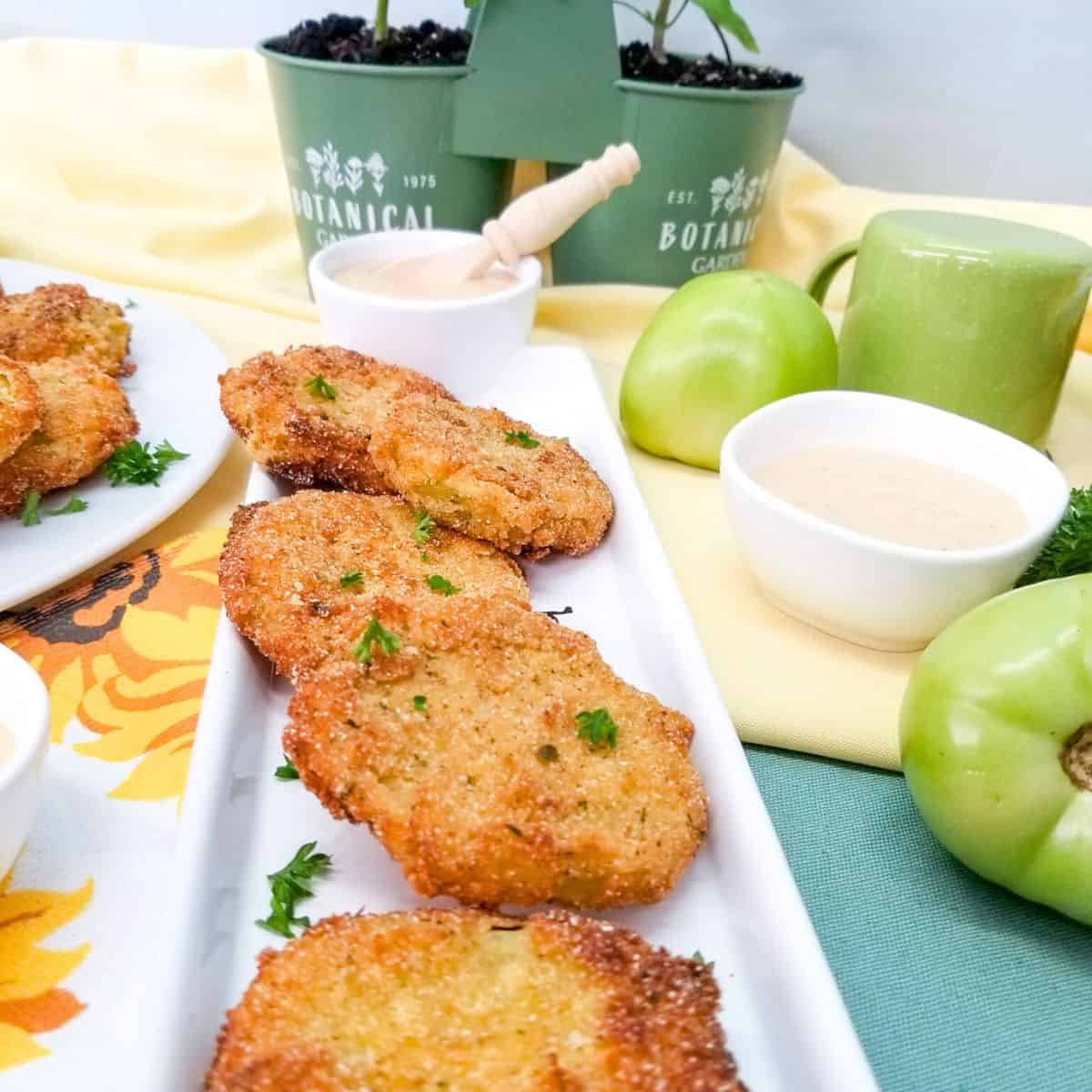 Fried tomatoes on a white plate with dipping sauce and whole green tomatoes in the photo.