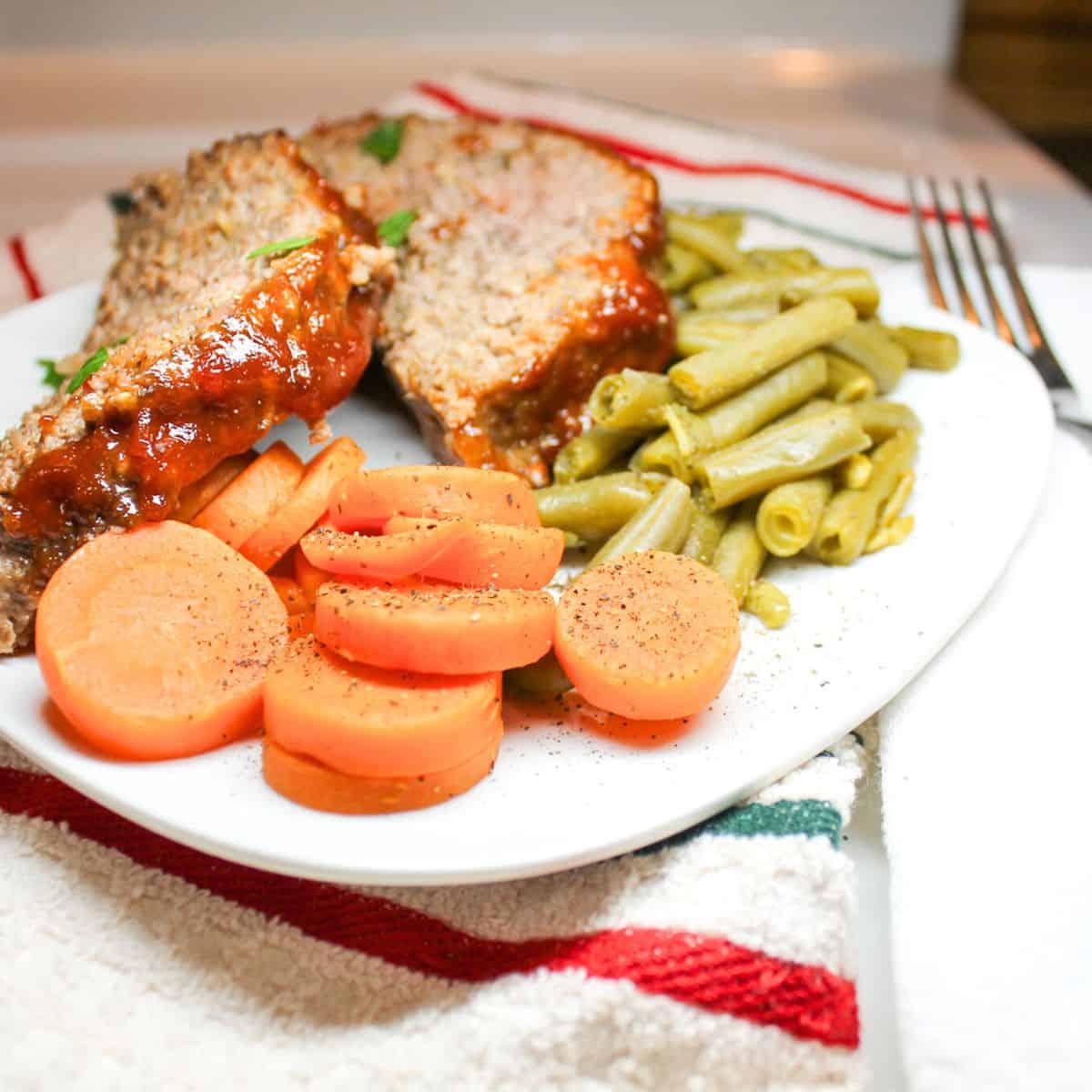 Sliced meatloaf on a white plate with green beans and carrots.
