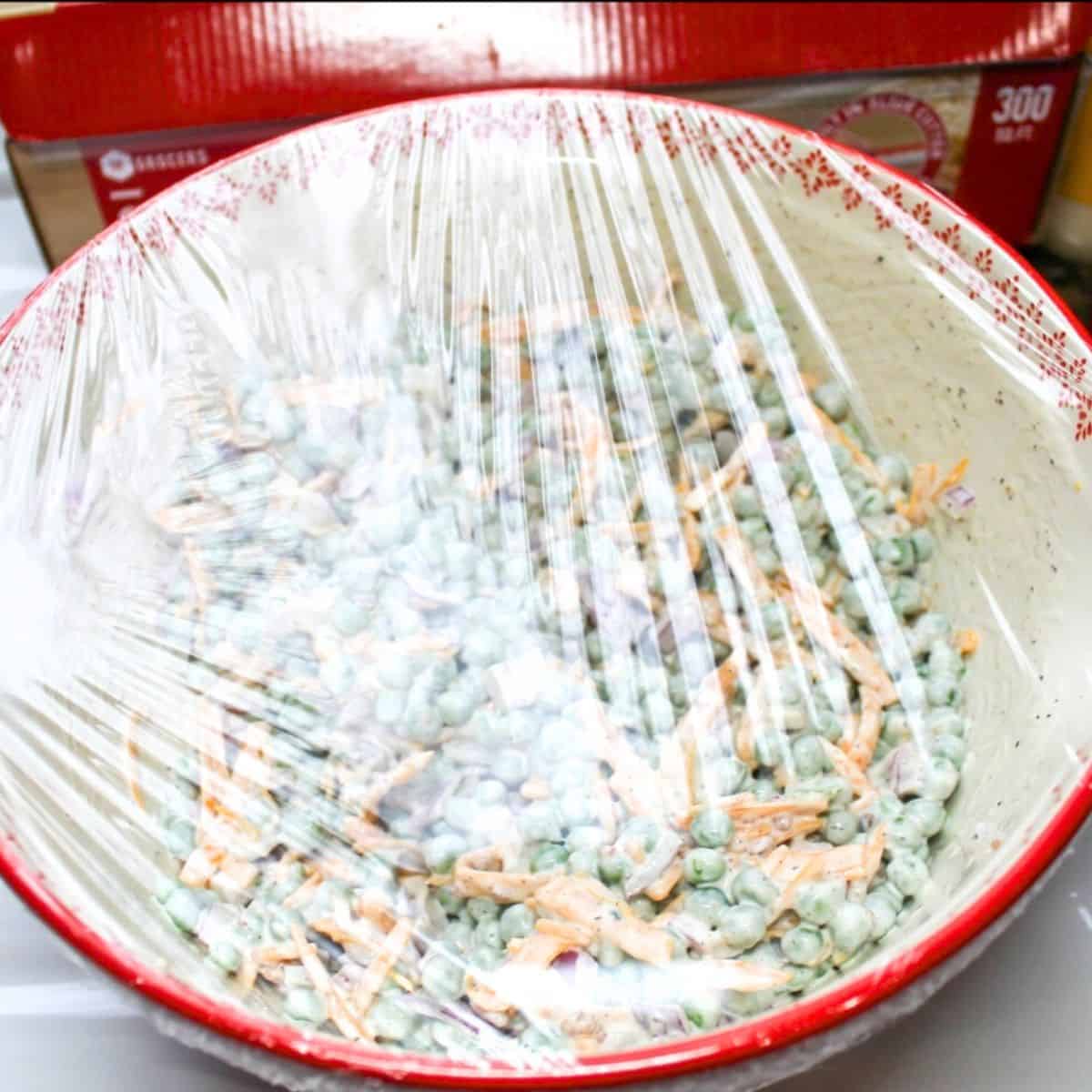 The pea salad with clear plastic wrap over a red bowl ready to be chilled in the refrigerator.
