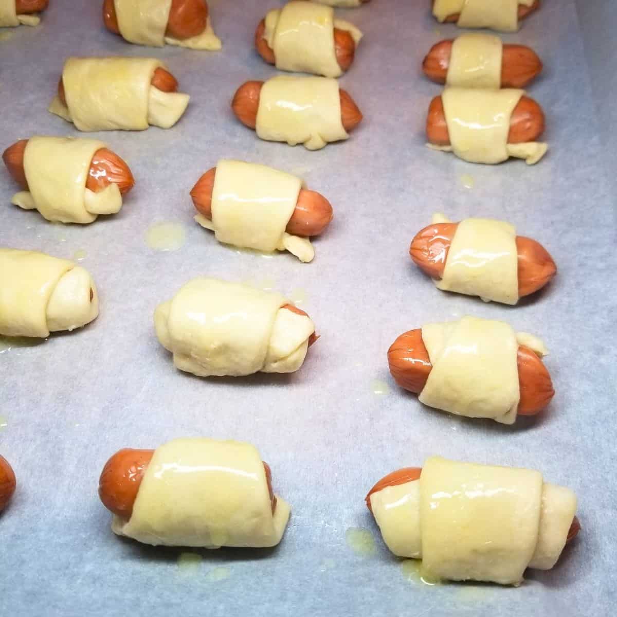 Pigs in a blanket with crescent rolls on a baking sheet covered with melted butter and ready for the oven.