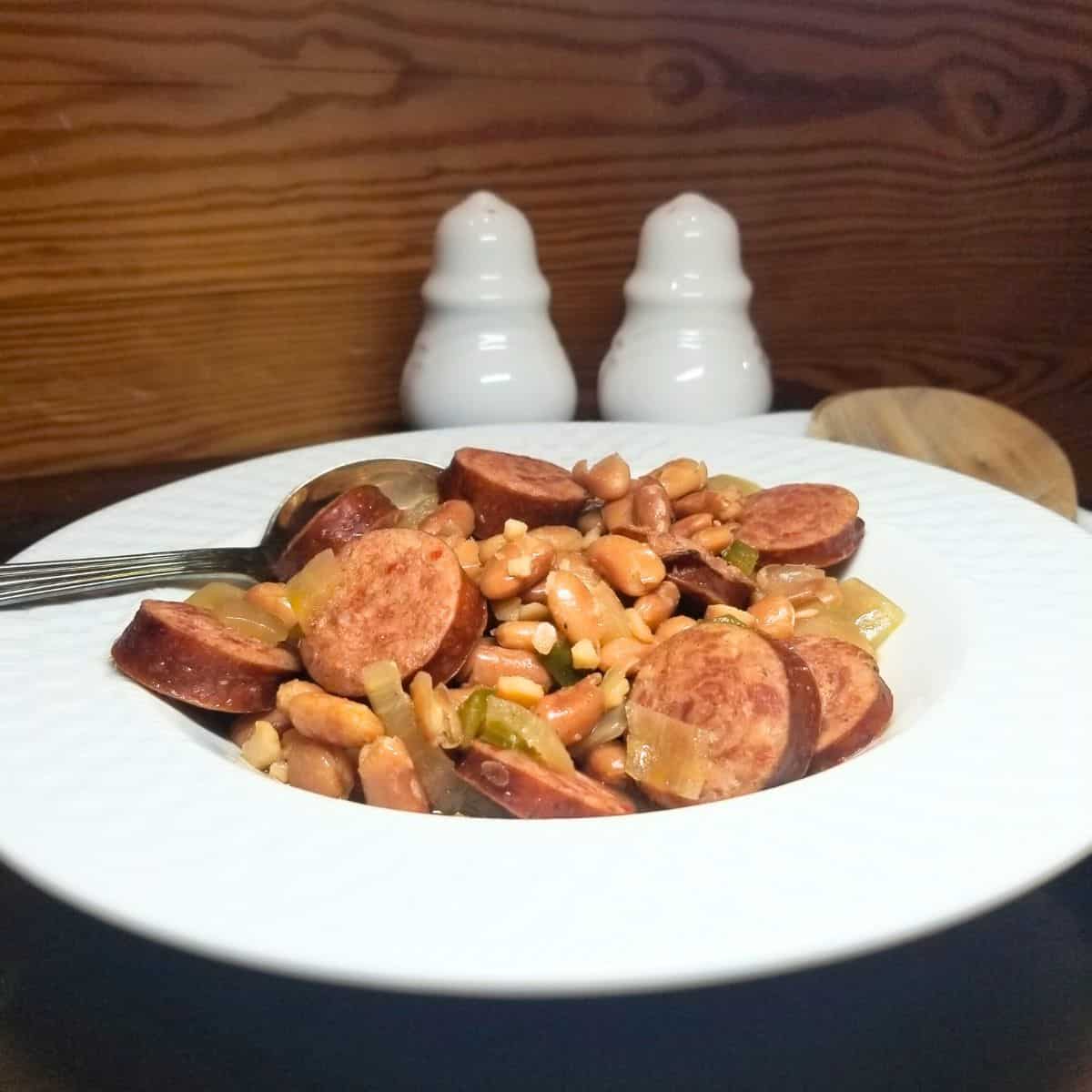 Bowl of slow cooker pinto beans and smoked sausage in a white bowl on a farmhouse table.