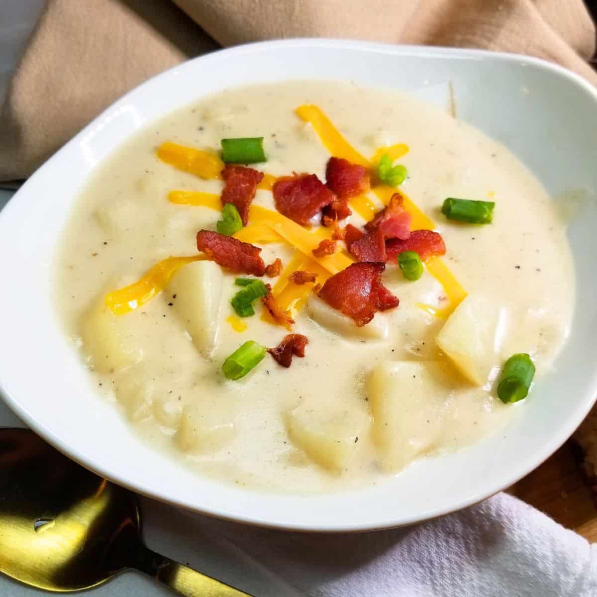 Potato soup in a white bowl topped with bacon, cheese, and green onions. A gold spoon and napkin next to the bowl.