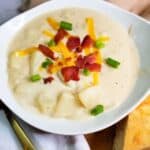 Potato Soup in a white bowl topped with bacon, cheese, and green onions. Gold spoon and cornbread.