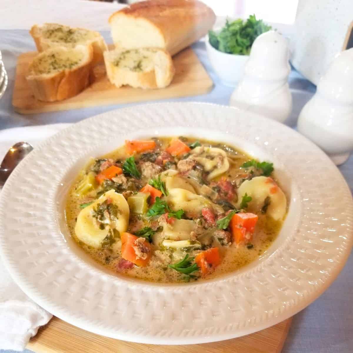 Sausage Tortellini soup in a white bowl with bread, parsley and shakers in the background.