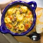 Tortellini soup in a blue bowl sitting on a wooden board with crusty bread and a spoon.