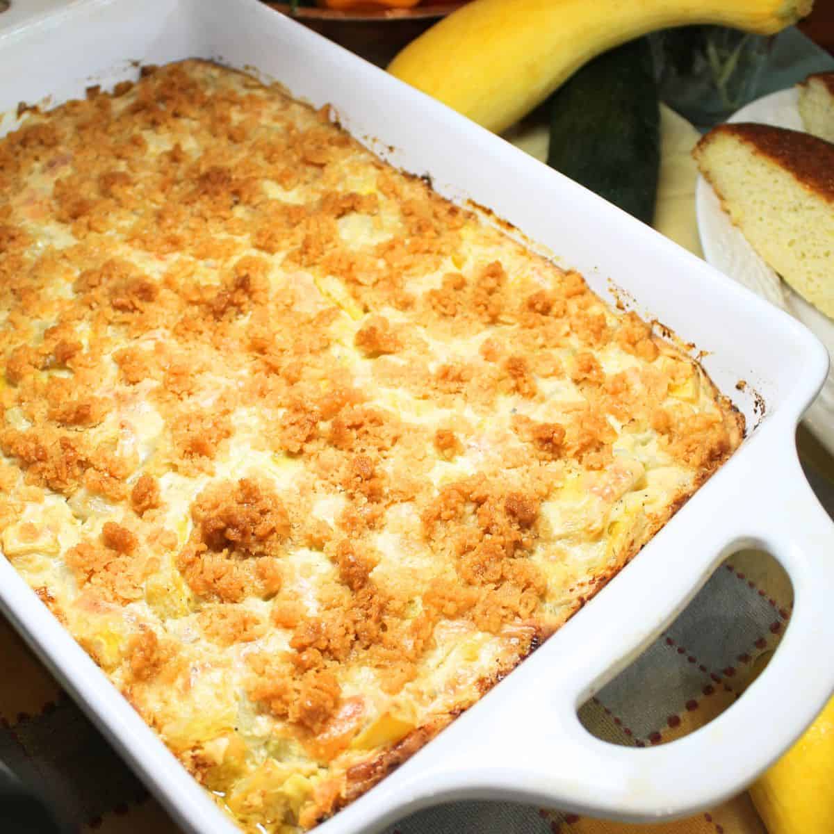 The baked squash casserole in a white baking dish. Yellow squash and cornbread in the background.