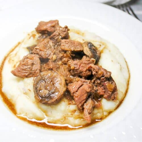 Steak bites with gravy over mashed potatoes in a white bowl with a fork.