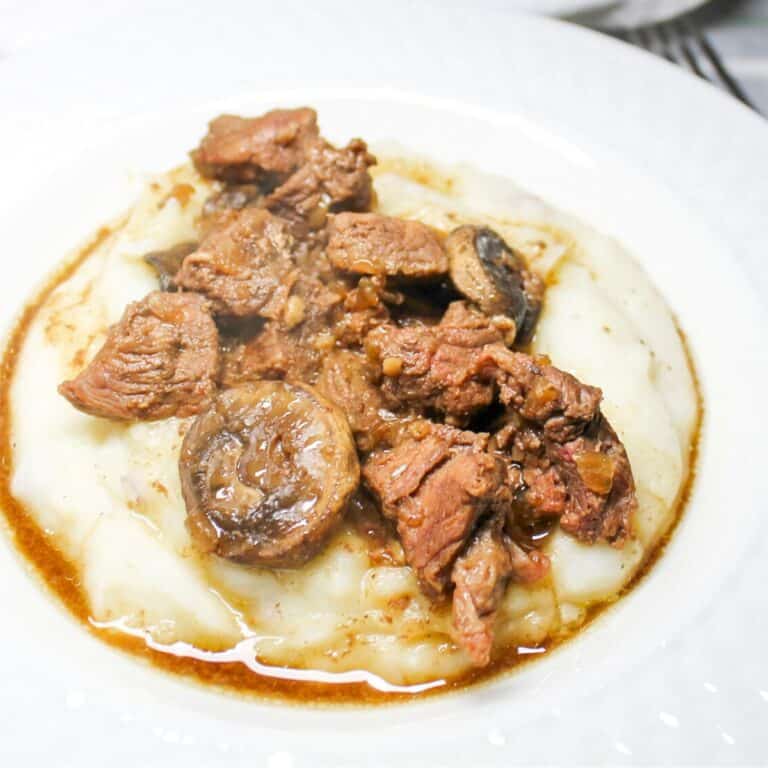 Steak bites with gravy over mashed potatoes in a white bowl with a fork.