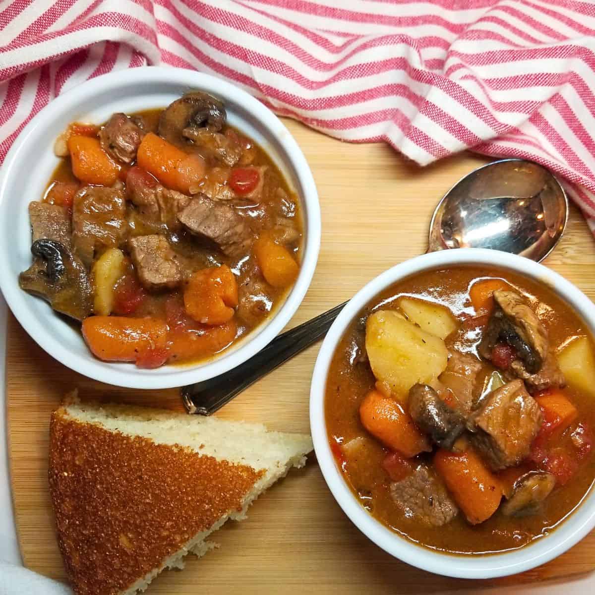 Two bowl of beef stew in white bowls with a spoon, cornbread, and stripped dish towel.