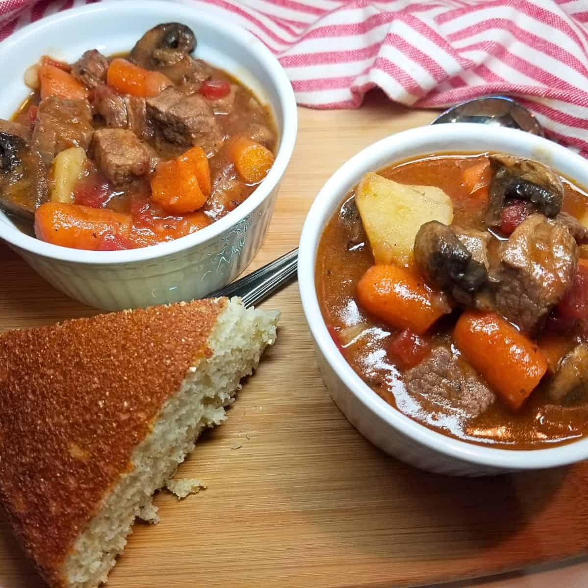 Two bowl of beef stew in white bowls with cornbread, a spoon, and stripped dish towel.