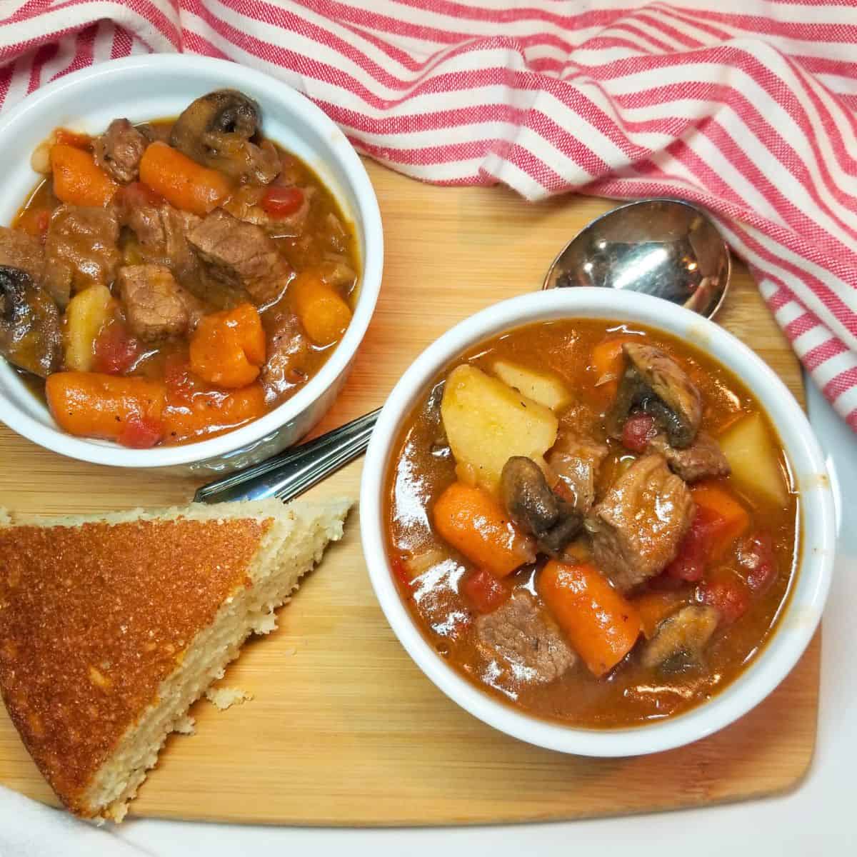 Stove top beef stew in white bowls with a spoon, cornbread and a red and white dish towel.