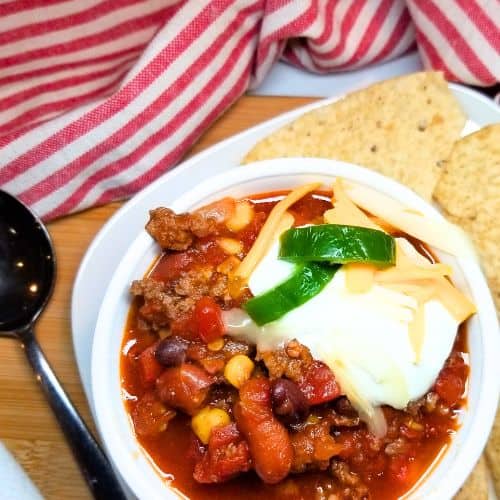 Soup in a white bowl with toppings, a spoon, tortilla chips and a dish towel in the background.