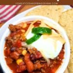 Soup in a white bowl with sour cream, cheese, and jalapenos on top. Tortilla chips and a dish towel on the side. Text overlay.