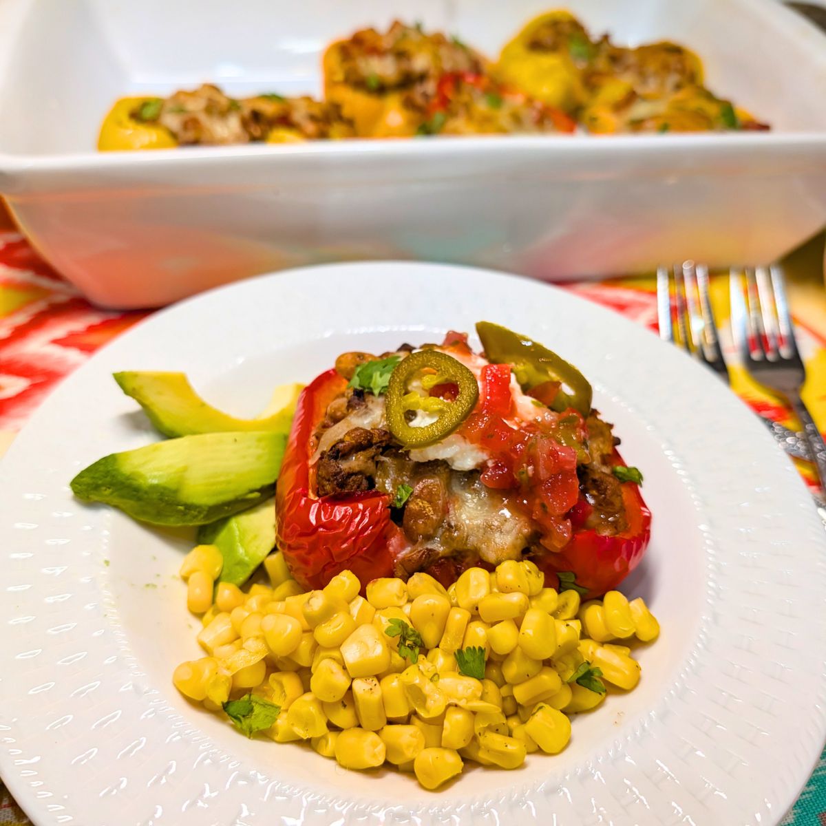 A stuffed red bell pepper, corn, and avocados on a white plate with a white baking dish in the background.