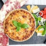 Baked tomato pie topped with basil. Some ingredients next to the pie with a kitchen towel.