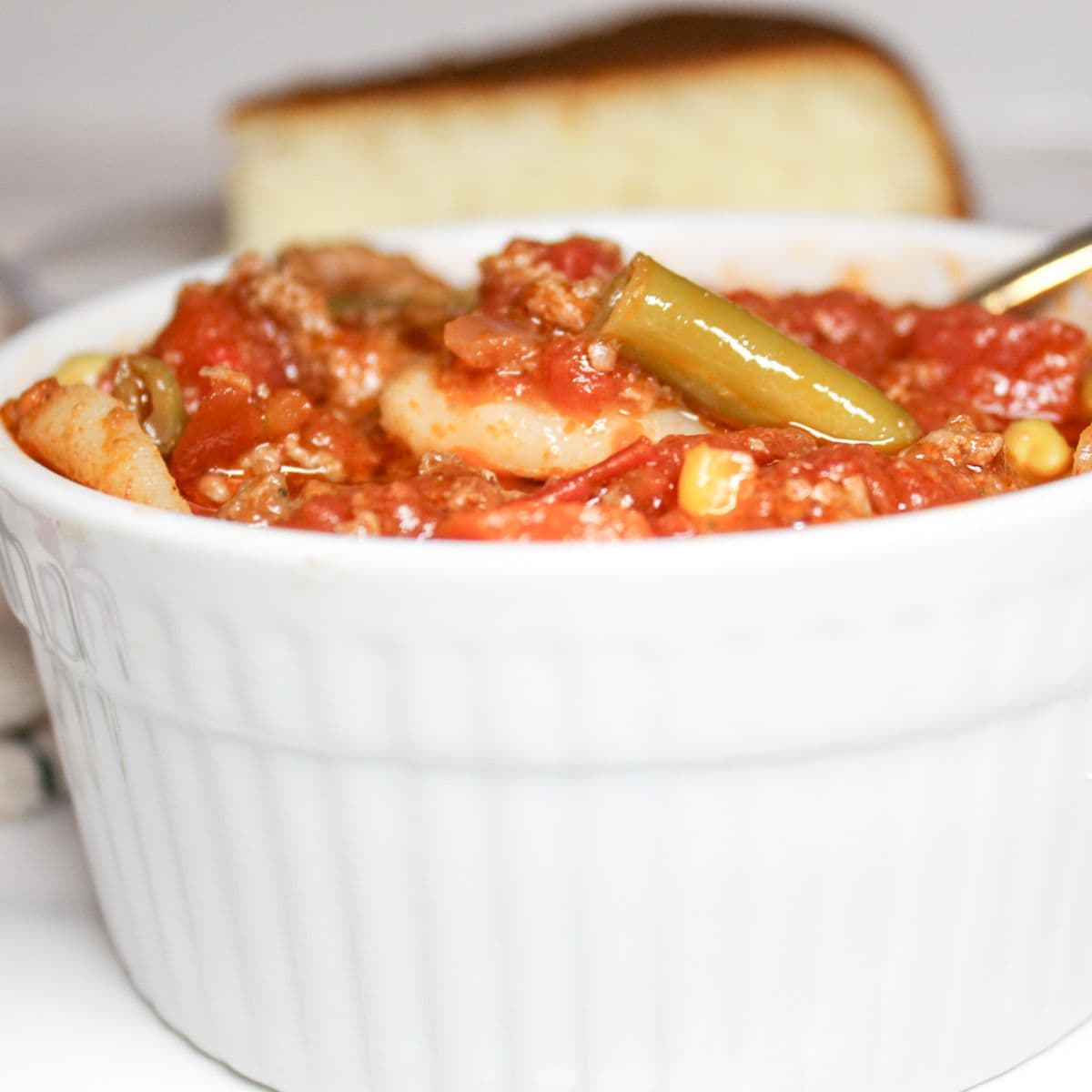 A white bowl of vegetable soup with a slice of cornbread in the background.