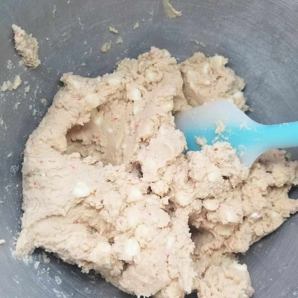 Cookie dough with white chocolate chips and peppermint in a mixing bowl with a blue spatula.