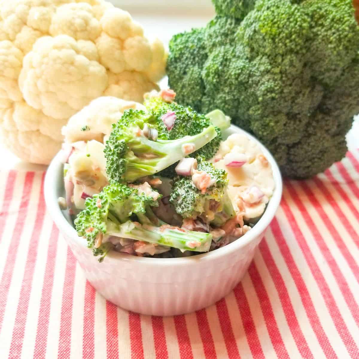 Salad in a white bowl with a head of cauliflower and broccoli in the background. Red and white stripped dish towel under bowl.